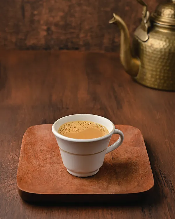 A white cup of milky tea on a wooden tray, with a brass kettle behind